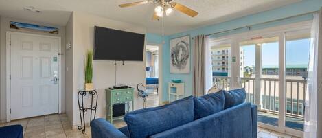 A living room with a blue sofa, a ceiling fan, a flat-screen TV, and a view of the ocean through the sliding glass doors.