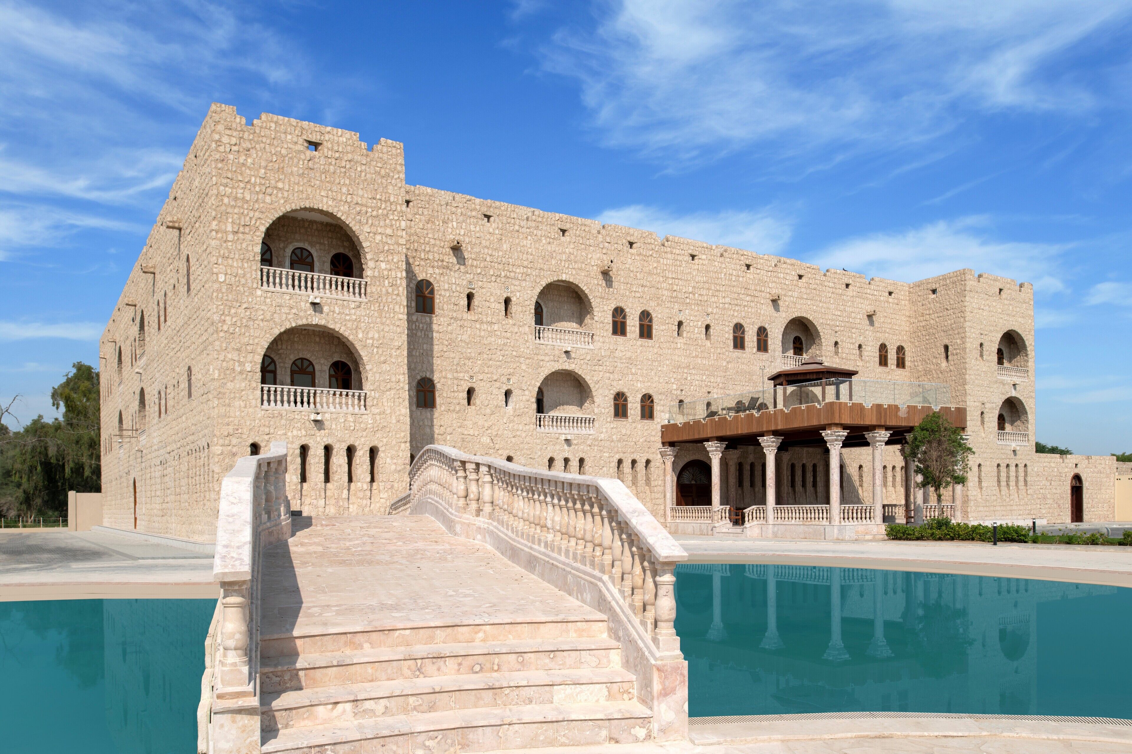 A large stone building with multiple arched windows and balconies, a swimming pool, and a grand staircase leading to the entrance.