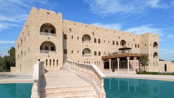 A large stone building with multiple arched windows and balconies, a swimming pool, and a grand staircase leading to the entrance.