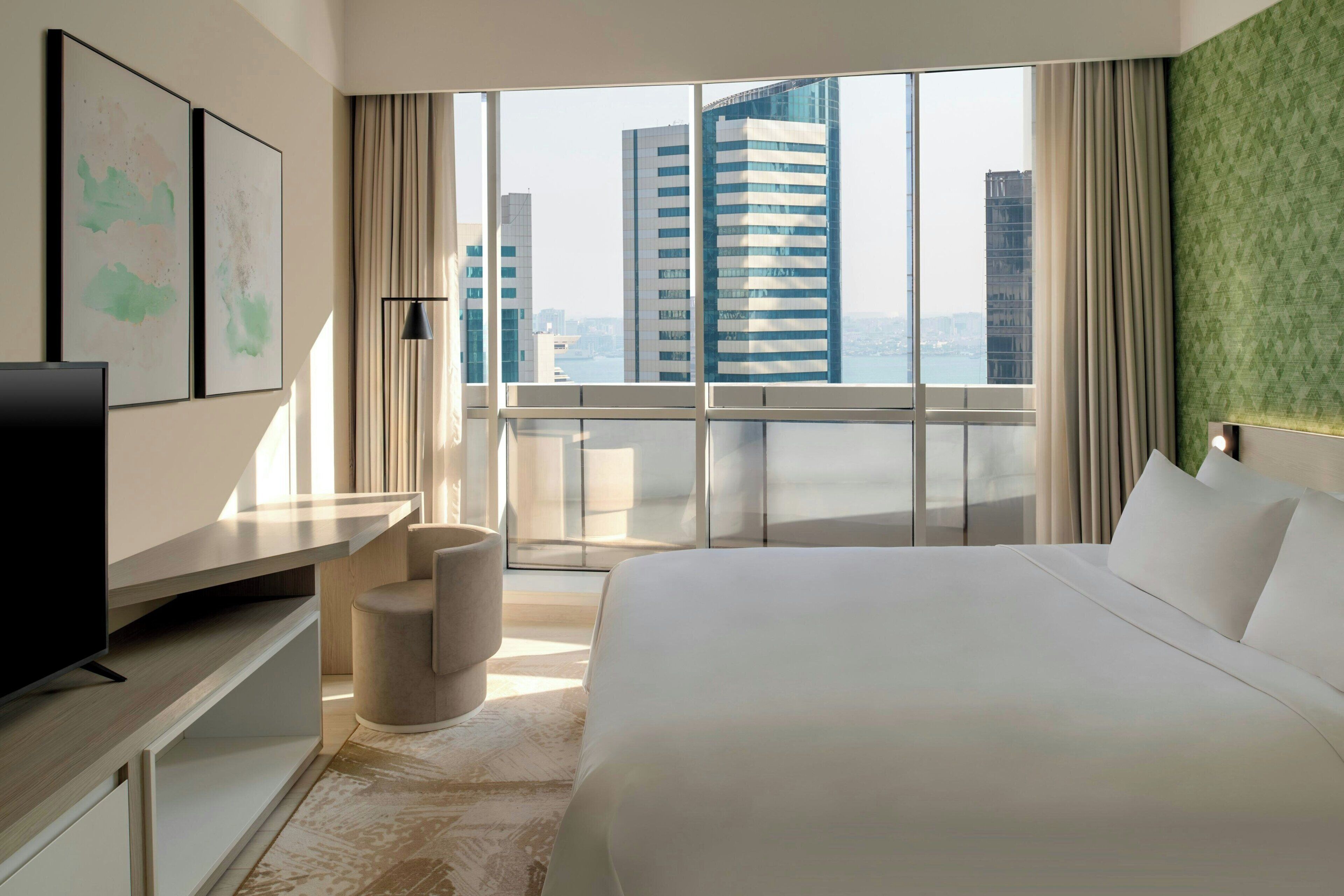 Habitación de hotel moderna con una cama grande, un escritorio, un televisor y vistas a altos edificios a través de amplios ventanales.