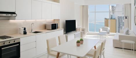 Une cuisine moderne avec des meubles blancs, un coin repas avec une table et des chaises blanches, un téléviseur à écran plat et une vue sur la mer grâce à de grandes fenêtres.