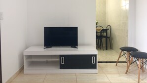 Una sala moderna con un televisor de pantalla plana sobre un mueble blanco, un comedor con mesa y sillas, y un baño visible a través de una puerta abierta.