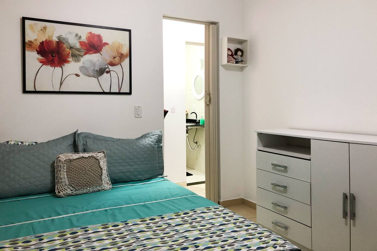 Quarto com cama, penteadeira, quadro e porta que dá para um banheiro.