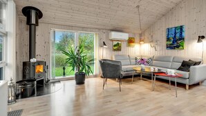 Een gezellige woonkamer met een houtkachel, een grijze bank, een salontafel en een groot raam met uitzicht op groen.