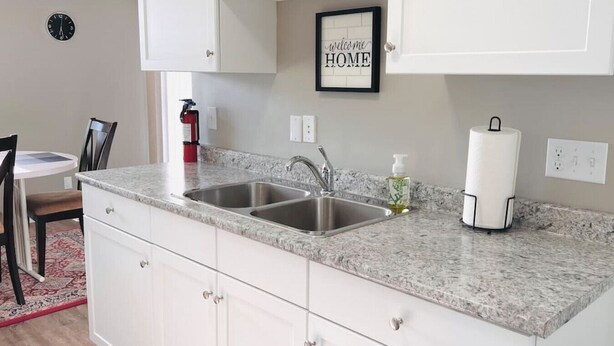 A kitchen with white cabinets, a double sink, a framed welcome sign, and a fire extinguisher. - Spacious 2-bedroom apartment (Camrose)