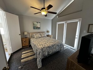 A bedroom with a bed, a ceiling fan, a nightstand, a lamp, a television, and a door. - Just steps to the Beach, Two Min Walking Distance (Galveston)
