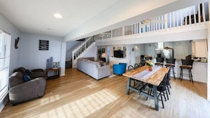 Uma sala ampla com piso de madeira, um sofá, uma mesa de jantar com cadeiras, uma escada e uma cozinha.