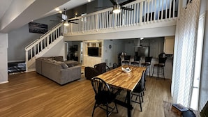 Ampla sala de estar com mesa de jantar de madeira, cadeiras pretas, sofá cinzento e escada para um andar superior.
