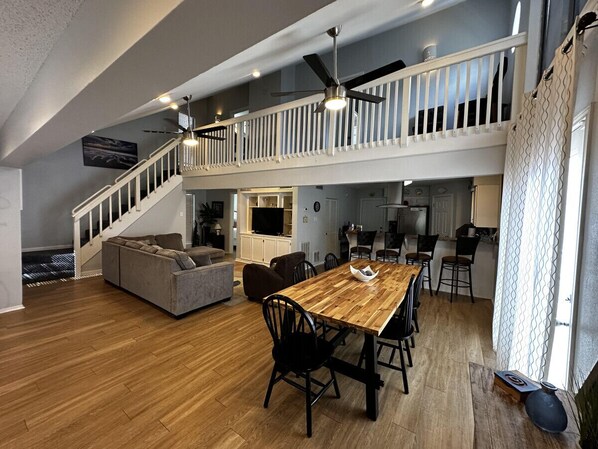 A spacious living area with a wooden dining table, black chairs, a grey sofa, and a staircase leading to an upper level.