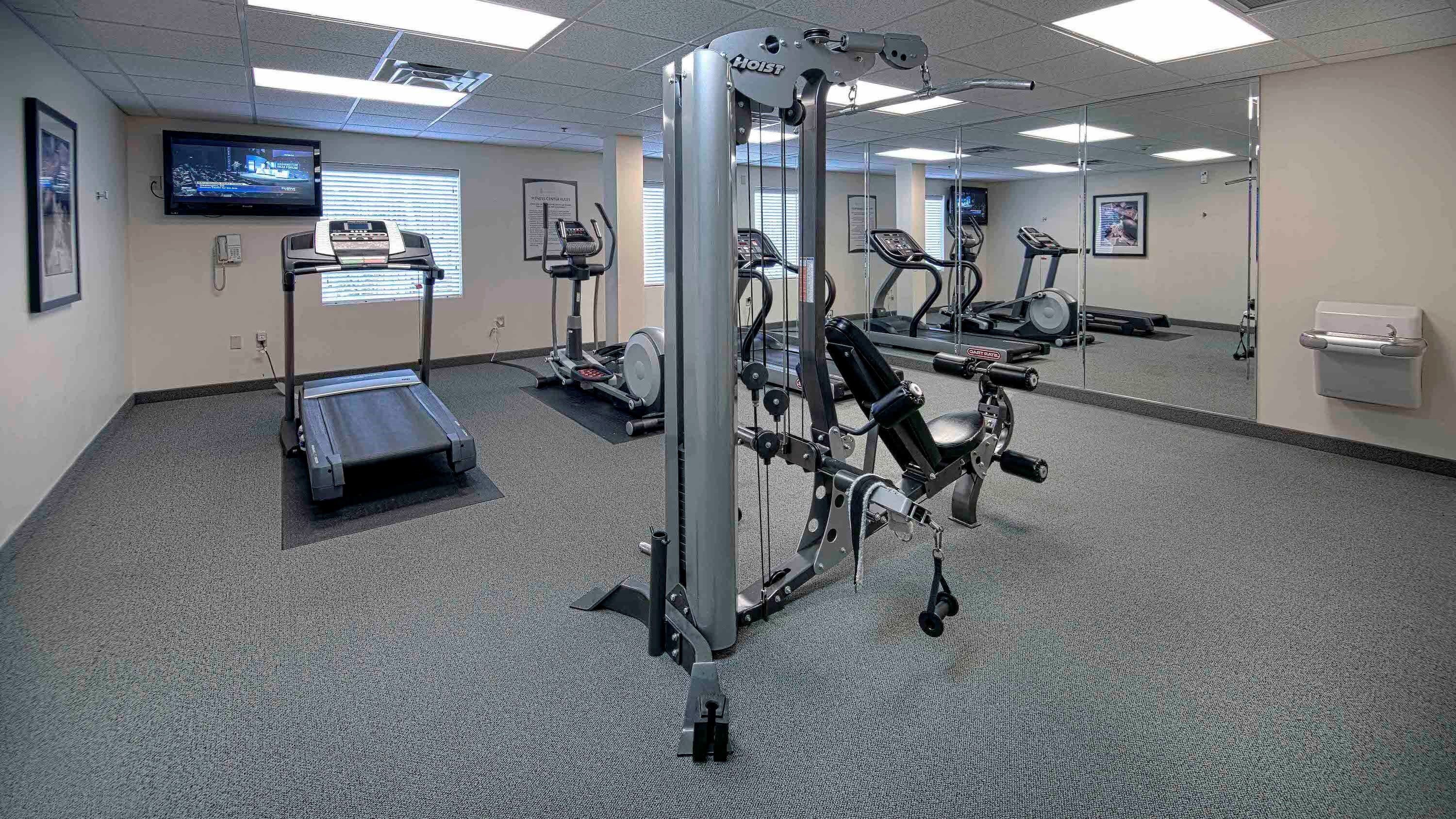 Un gimnasio con máquinas de cardio, equipo de pesas y una televisión que muestra noticias.