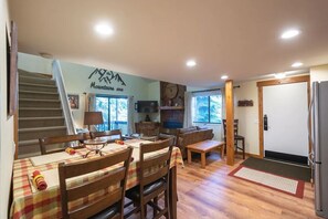 A dining area with a table set for four, a sofa, a TV, and a staircase leading to another level.