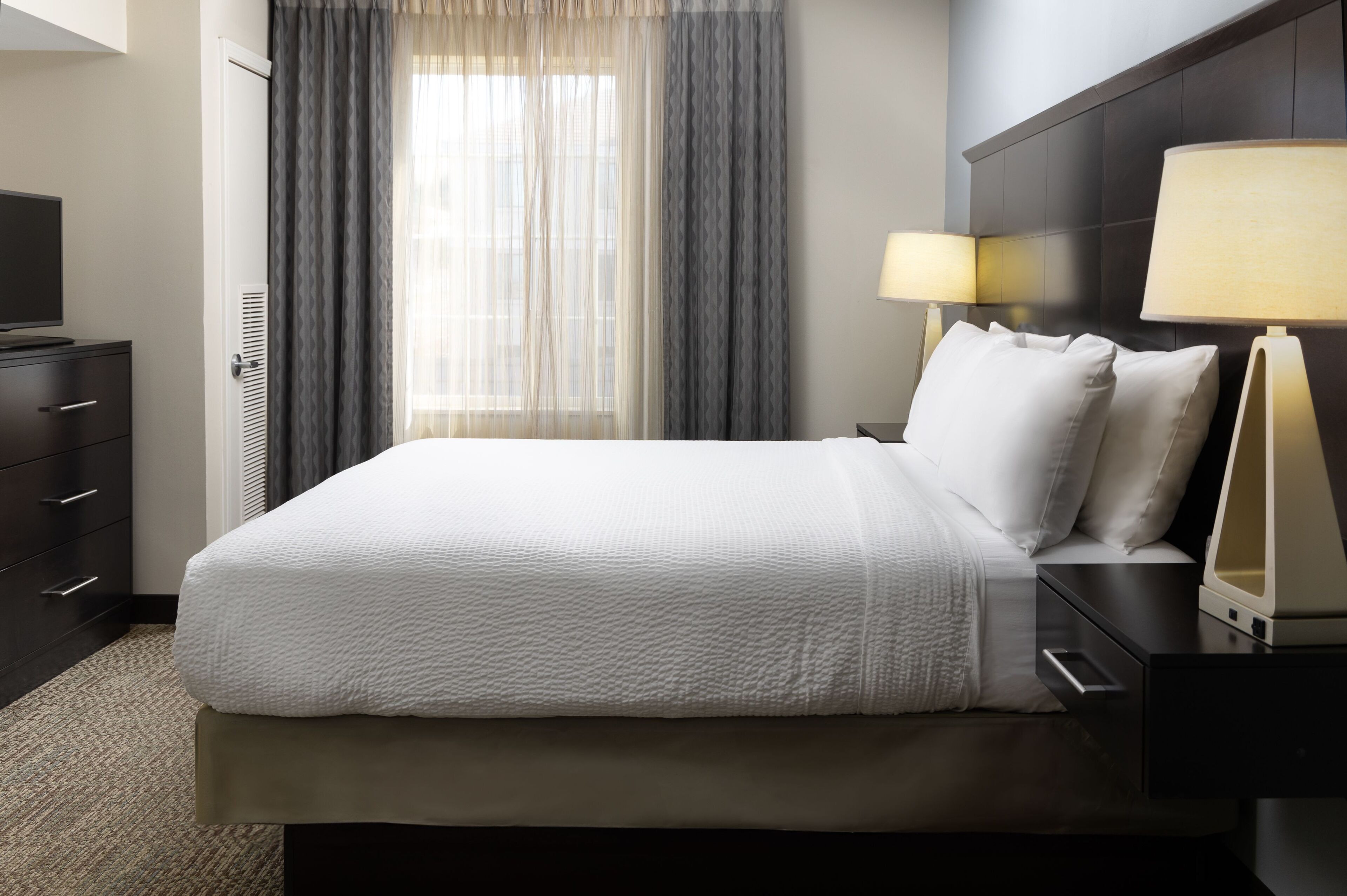 A hotel room with a bed, nightstands, a TV, a dresser, and a window with curtains.