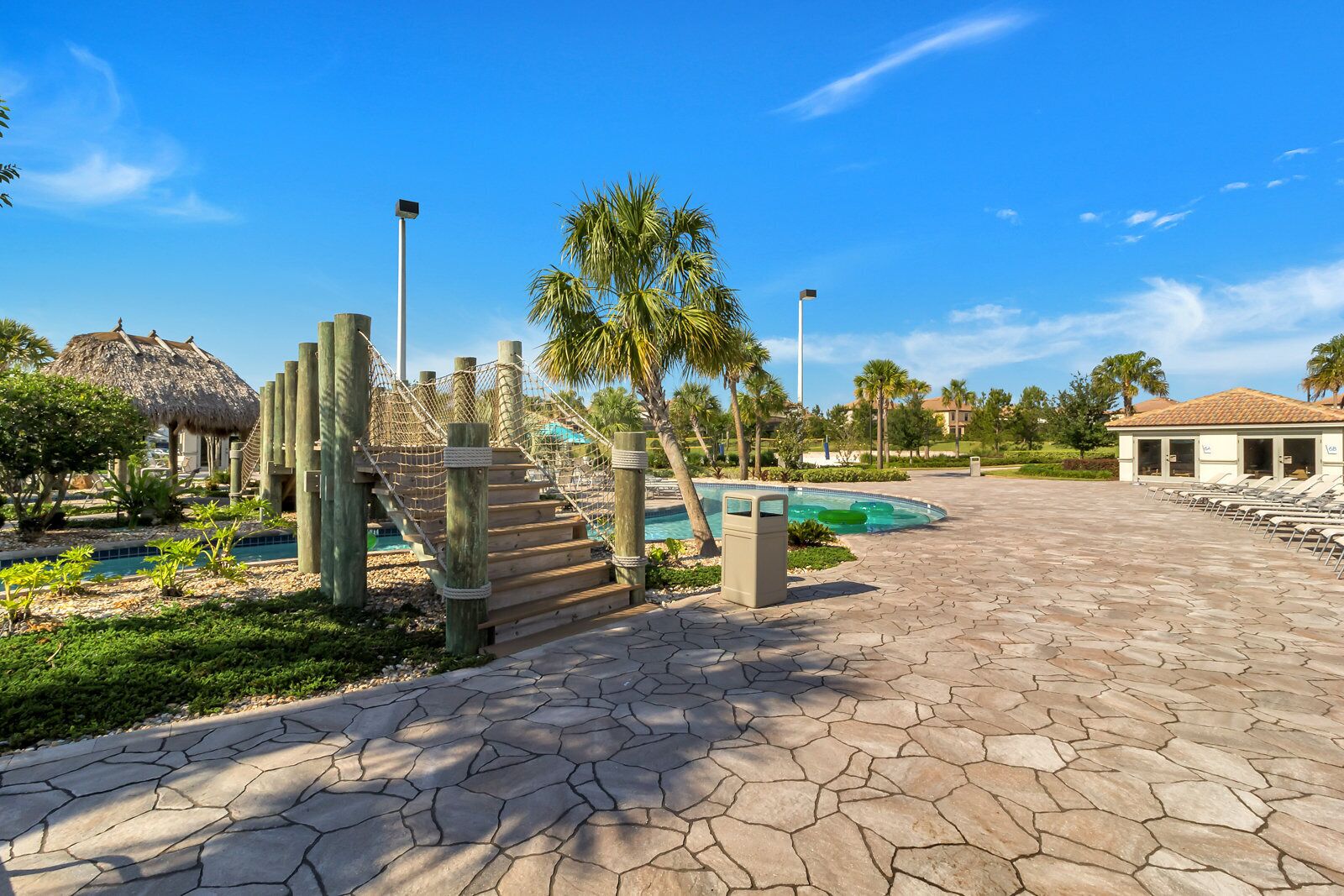 A pool area with a wooden staircase, palm trees, and a thatched-roof structure.
