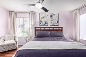 Un dormitorio con una cama, una silla, un ventilador de techo y dos cuadros abstractos en la pared.