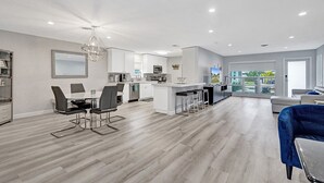 A modern kitchen with white cabinets, a dining area with a glass table, and a living room with a blue sofa.