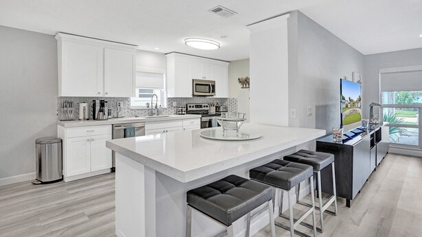 A modern kitchen with white cabinetry, a central island with stools, stainless steel appliances, and a large window providing natural light.
