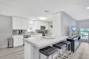 A modern kitchen with white cabinetry, a central island with stools, stainless steel appliances, and a large window providing natural light.