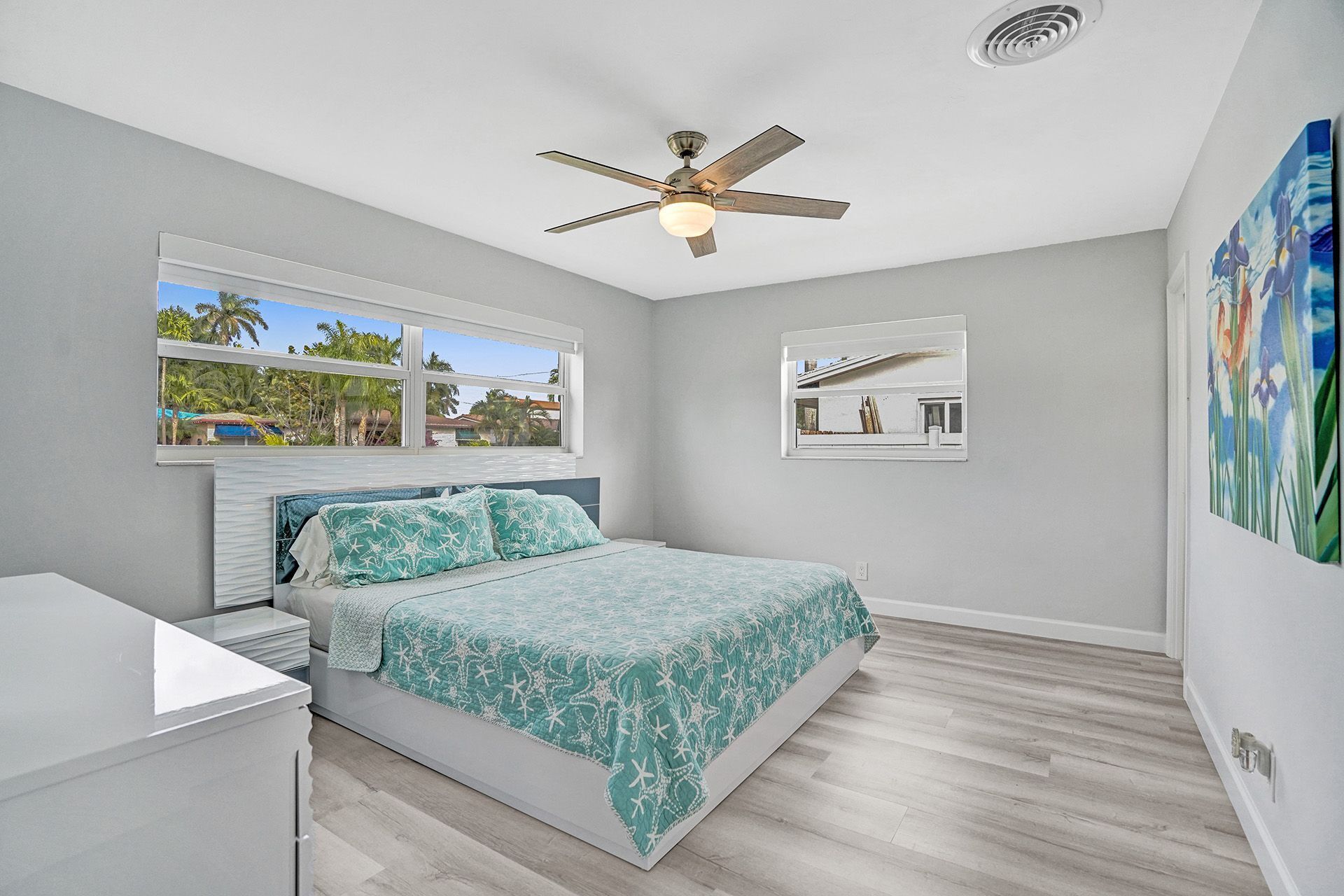 A bedroom with a bed, a ceiling fan, a window with a view of palm trees, and a painting on the wall.