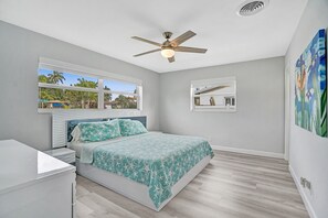 A bedroom with a bed, a ceiling fan, a window with a view of palm trees, and a painting on the wall.