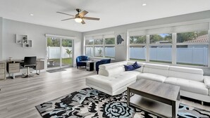 A spacious living room with a white sofa, a coffee table, and a desk with a chair. There are large windows offering a view of the backyard.