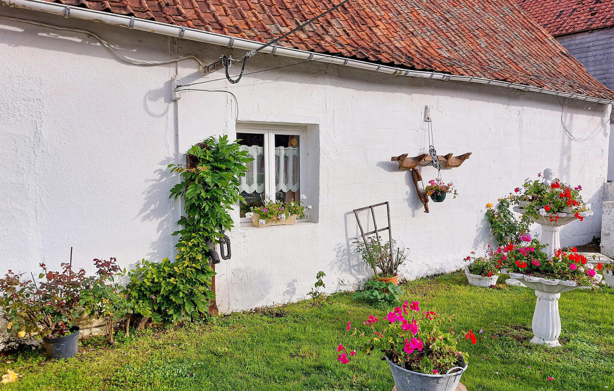 Um edifício branco com telhado de telhas, janela com persianas e vários vasos com plantas e flores no jardim.