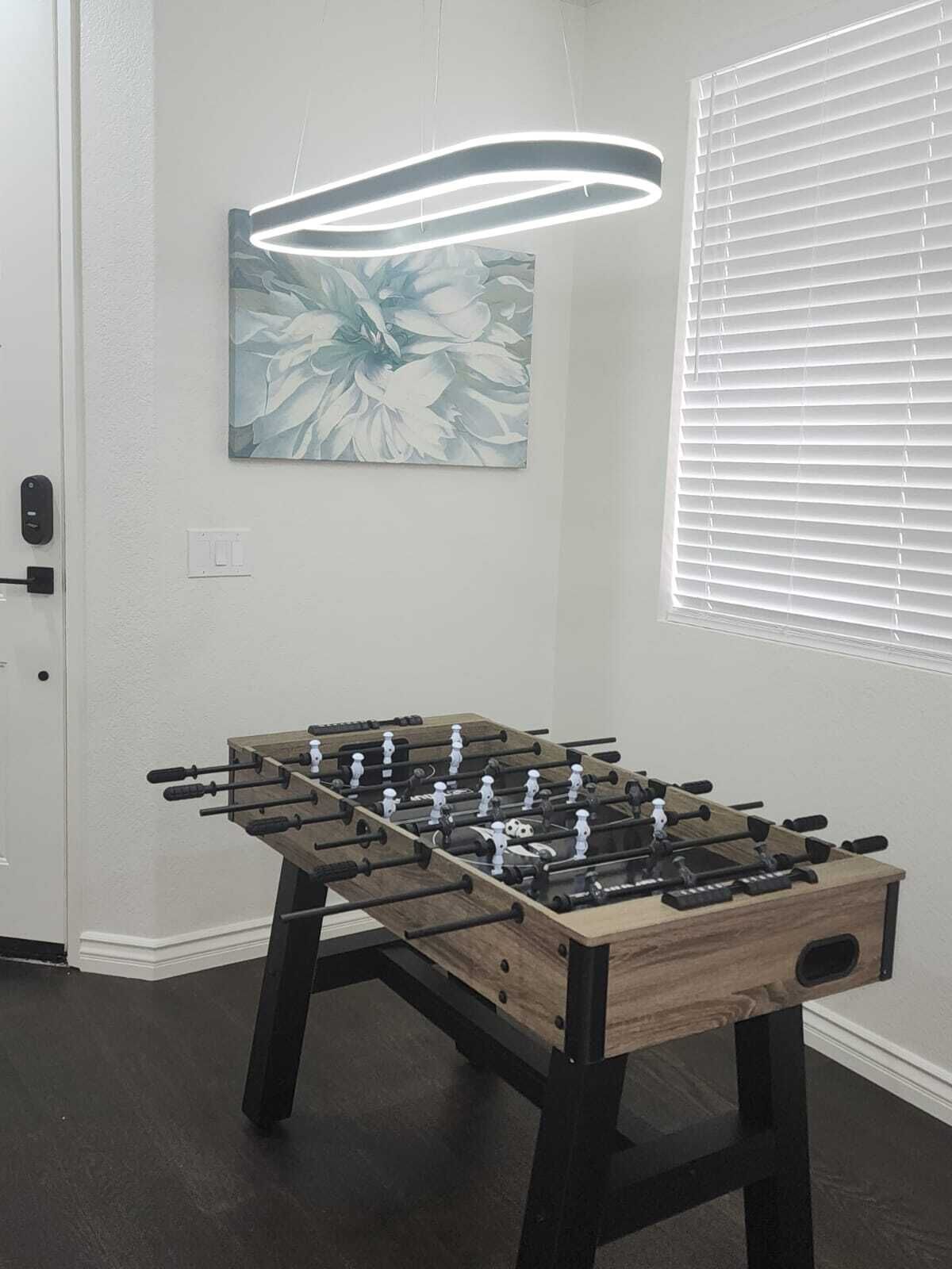 Una mesa de fútbolín en una habitación con una puerta blanca, un cuadro y una ventana con persianas.