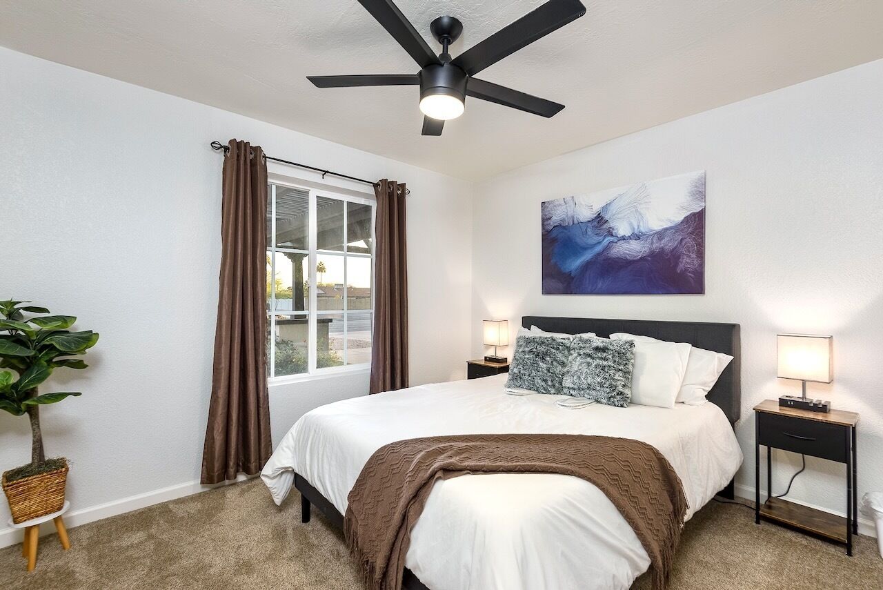 Ein Schlafzimmer mit Bett, Deckenventilator, Fenster mit Vorhängen, einem Wandbild und einem Nachttisch mit Lampe.