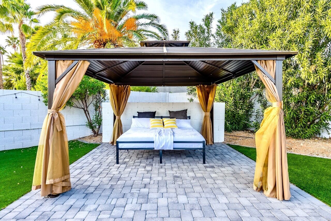 A bed under a pergola with curtains, surrounded by a brick patio and greenery.