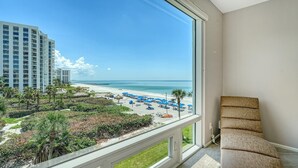 Une pièce dotée d’une grande fenêtre donnant sur une plage, d’un fauteuil et offrant une vue sur un bâtiment d’hôtel.
