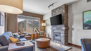 A modern living room with a stone fireplace, a large flat-screen TV mounted above it, and a comfortable seating area with a sofa and armchairs. - 3 Sisters Mountain Views | Hot Tub & Heated Pool (Canmore)