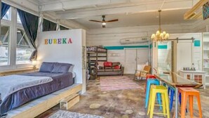 Une pièce avec un lit, un bureau doté de tabourets colorés, un ventilateur de plafond et un mur sur lequel est écrit le mot «EURÉKA».