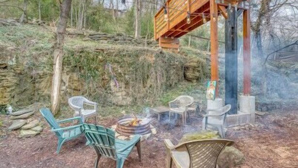 Un espace extérieur rustique aménagé avec des chaises et un foyer, entouré d’arbres et d’une structure en bois.