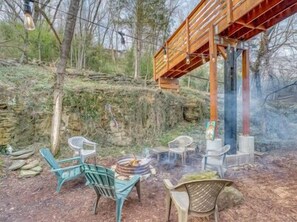 A rustic outdoor seating area with chairs and a fire pit, surrounded by trees and a wooden structure.
