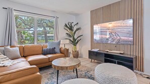 En moderne stue med en lædersofa, et rundt sofabord, et tv monteret på en væg af træpaneler, og et stort vindue med udsigt til grøn natur.
