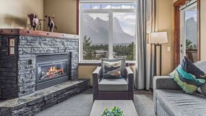 Una sala de estar con chimenea de piedra, un sofá, un sillón y vistas a montañas a través de un ventanal.
