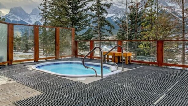 Un jacuzzi con vista a montañas nevadas, rodeado de barandales de madera y un banco de madera.