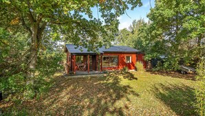 A small, red wooden cabin with a porch and a covered outdoor seating area, surrounded by trees and a grassy yard. - Amazing home in Rødby with WiFi (Rødby)