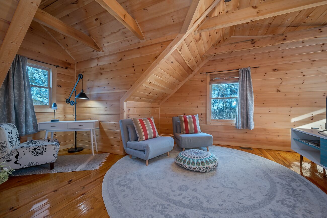 A cozy log cabin room with a wooden ceiling, two sofas with patterned cushions, a round rug, and a desk with a lamp.