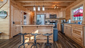Une cuisine rustique aménagée dans une cabane en bois, avec un îlot central, des tabourets de bar, des appareils en acier inoxydable et une grande fenêtre agrémentée d’un parasol.