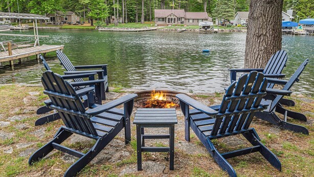 Un paisaje con lago, una fogata y sillas Adirondack.