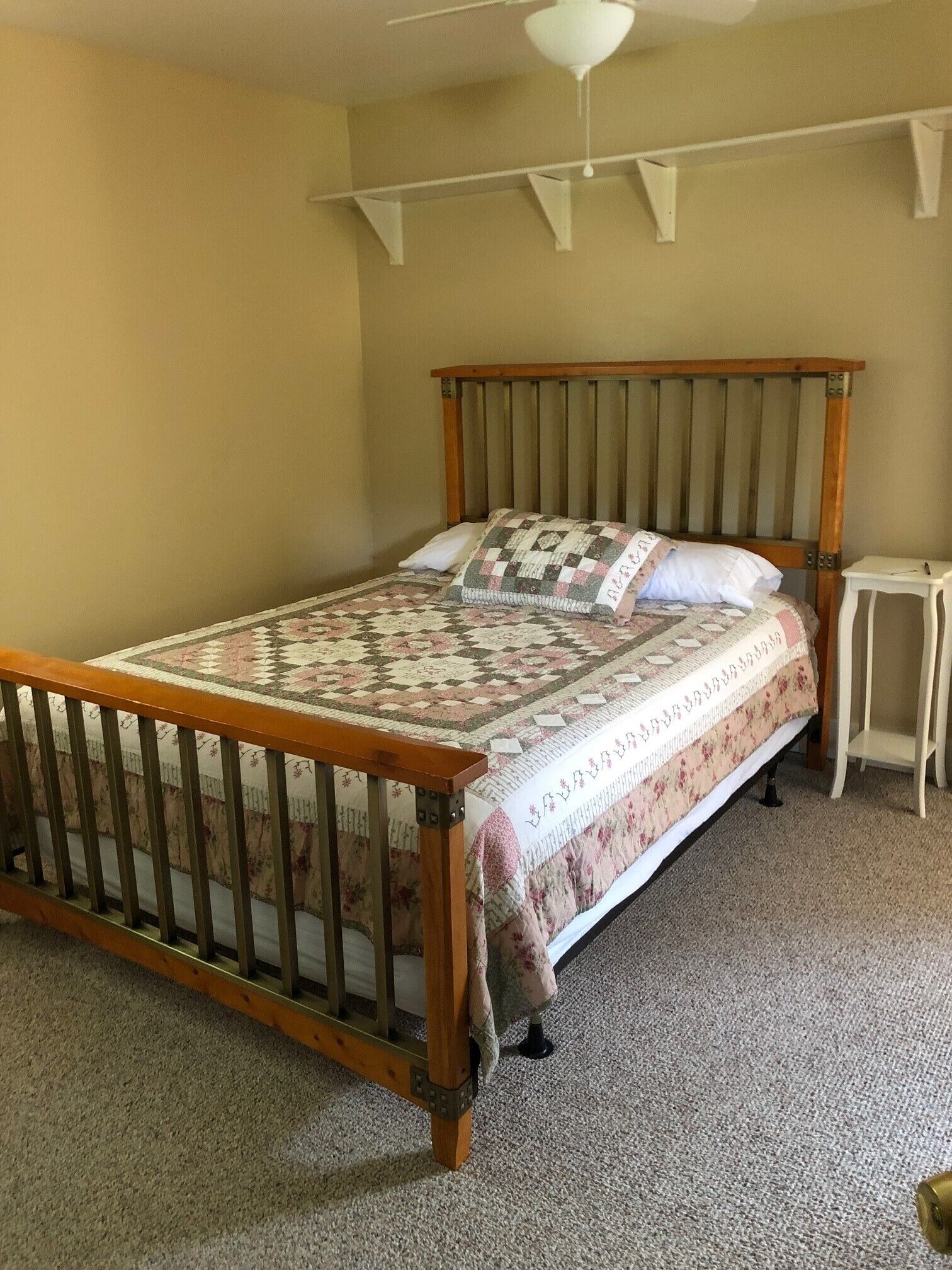 Quarto com estrutura de cama de madeira, colcha, mesa de cabeceira pequena e ventilador de teto.