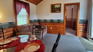 A cozy room with a wooden ceiling, a sofa, a dining table set with plates and glasses, and a painting on the wall. - Breezy Meadow Two 2 of 2,custom built cottage, under a mile from CDP (Milford)