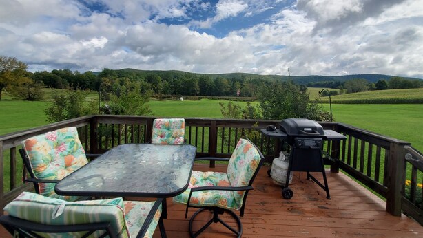 A wooden deck with a table and chairs, a grill, and a view of a green field and hills. - Breezy Meadow Two 2 of 2,custom built cottage, under a mile from CDP (Milford)