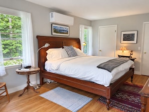 A bedroom with a wooden bed, a bedside table with a lamp, a window with curtains, and a door. - Comfortable Country Charm with Otsego Lake Access (Cooperstown)