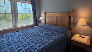 Ein Schlafzimmer mit großem Fenster, einem Holz-Kopfteil, einer Nachttischlampe und einem Bett mit einer blauen, strukturierten Decke.