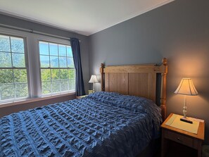 A bedroom with a large window, a wooden headboard, a bedside lamp, and a bed with a textured blue blanket. - Changing Seasons Game room, large yard, 3 bedroom/2 bath ranch, country setting (Hartwick)