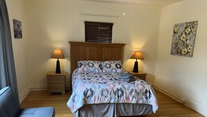A bedroom with a bed, two nightstands, a lamp on each, a wooden headboard, a window with blinds, a couch, and a floral painting on the wall. - City Fox 1, 1 of 4, recent renovation in Oneonta, Convenient to All Star Village (Oneonta)