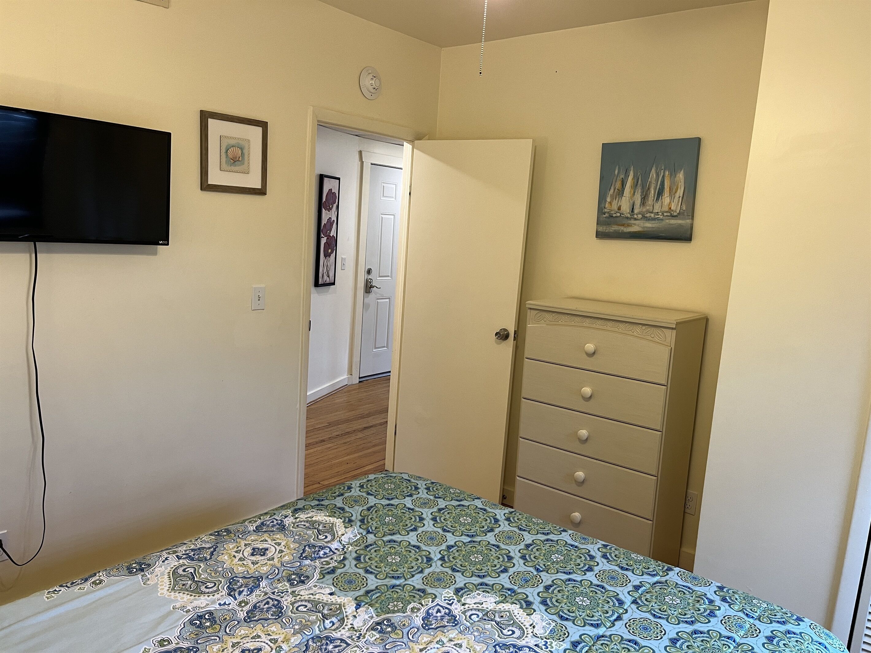 Ein Schlafzimmer mit einem geblümten Bettbezug, einem Schminktisch, einem an der Wand befestigten Fernseher und einer Tür zu einem weiteren Raum.