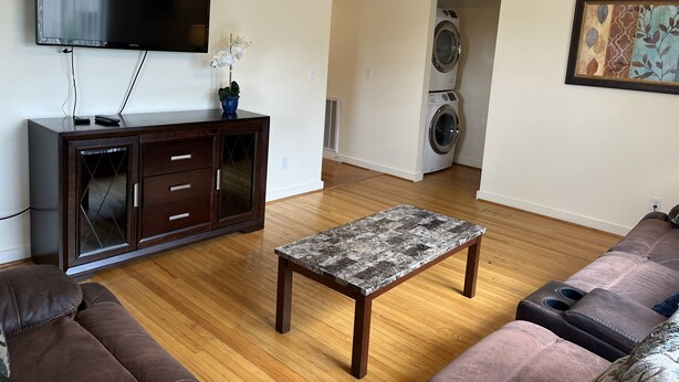Een woonkamer met een flatscreen televisie, een houten kast, een marmeren salontafel, een bank en een wasmachine op de achtergrond.