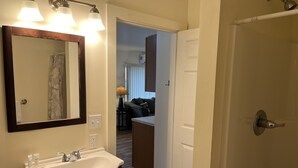 Une salle de bain avec un lavabo blanc, des meubles sombres et un miroir avec éclairage intégré. Une porte donne sur un espace de vie comprenant un canapé et une lampe.